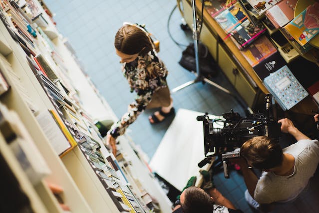 Operadores de câmara a filmar um vídeo cinematográfico de uma mulher a tocar em livros numa biblioteca.