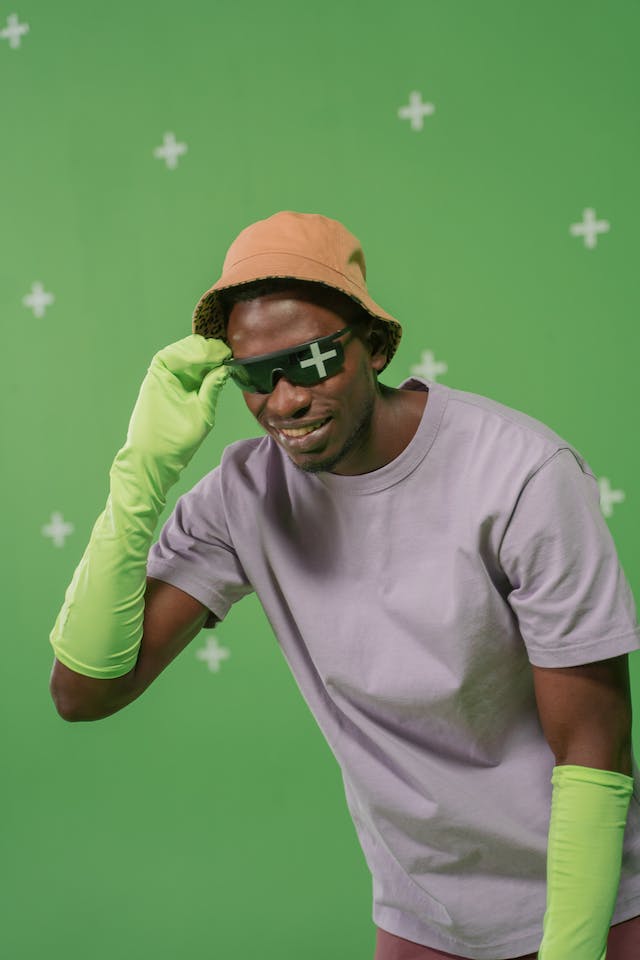 Un bărbat cu ochelari de soare și o pălărie de găleată pozând cu un ecran verde în spatele lui.