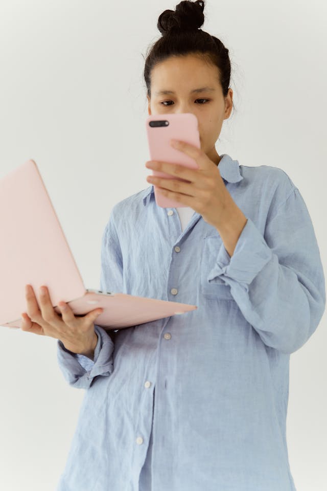 Une femme en chemise bleue tient un ordinateur portable et un téléphone. 