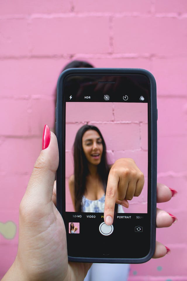 Cineva fotografiază o femeie cu telefonul, în timp ce aceasta pare că își întinde mâna în afara ecranului camerei foto, apăsând pe butonul de înregistrare
