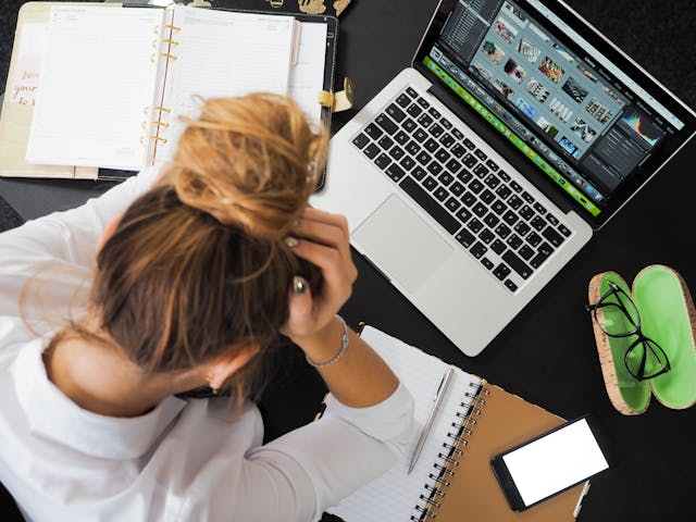 Een gestreste vrouw kijkt naar een laptop en notitieblokken terwijl ze haar hand op haar hoofd laat rusten.