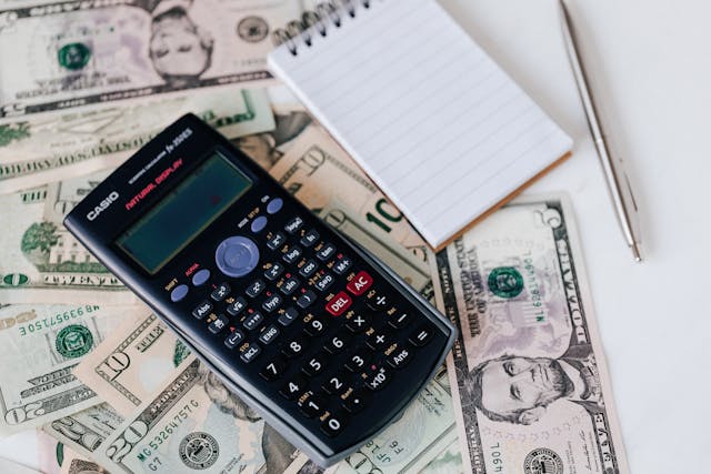 Uma calculadora, um bloco de notas e uma caneta sobre uma mesa coberta de notas de dólar americano.