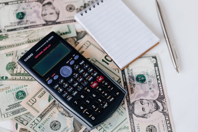 A calculator, notepad, and pen on a table covered in US dollar bills.