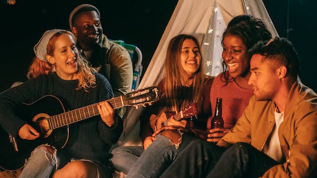 Un grup de prieteni care beau și cântă în timp ce sunt adunați noaptea într-un camping.