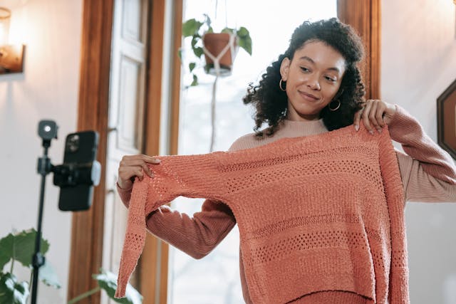 A woman filming herself while showing the fine details of a sweater.