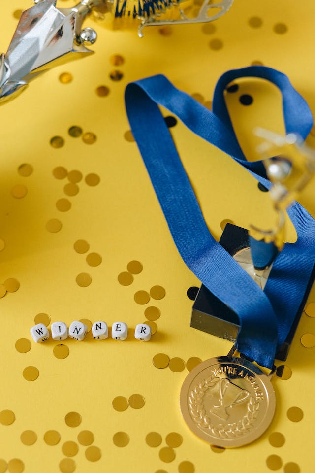 A gold medal on a yellow surface next to gold confetti and letter beads that spell the word “WINNER.”