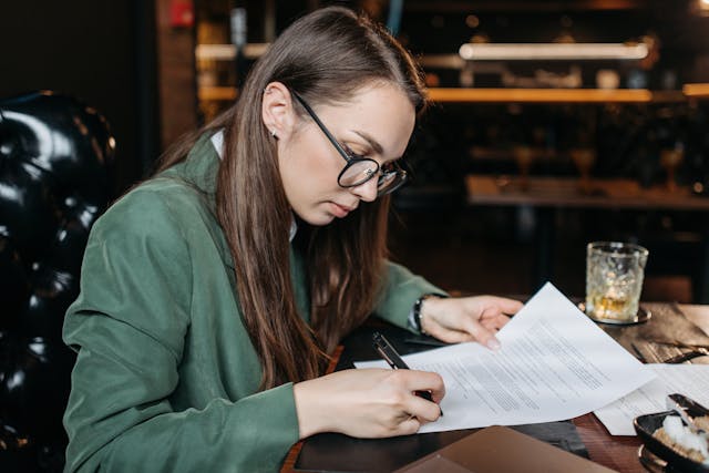 안경을 쓴 여성이 계약서를 읽고 서명하고 있습니다.
