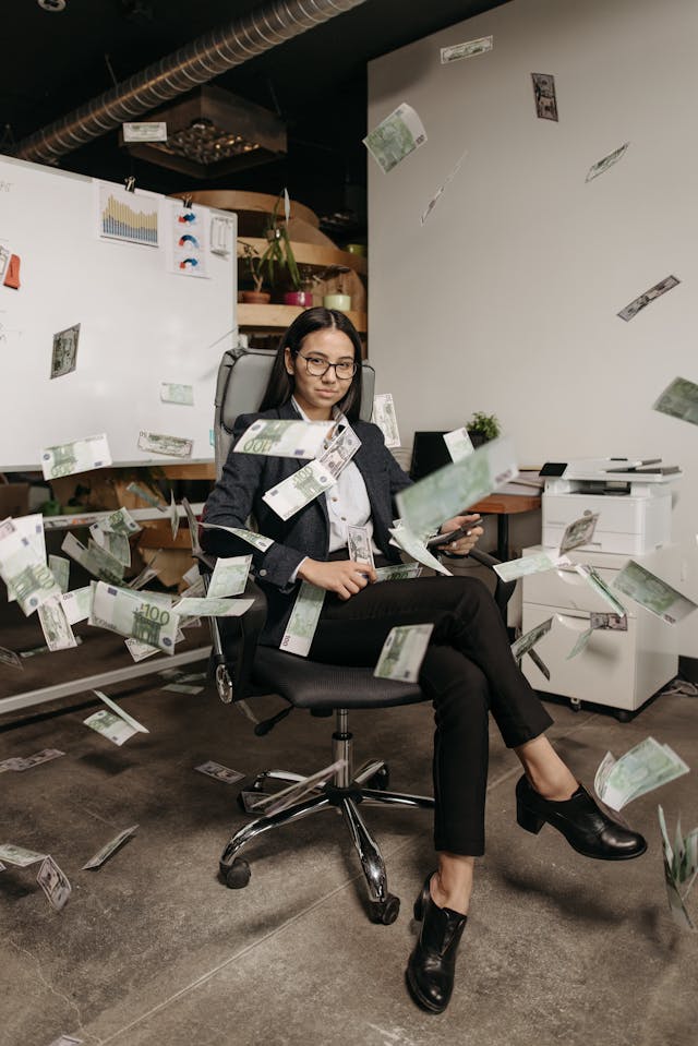 A young woman sitting in an office chair with dollar bills flying around her.