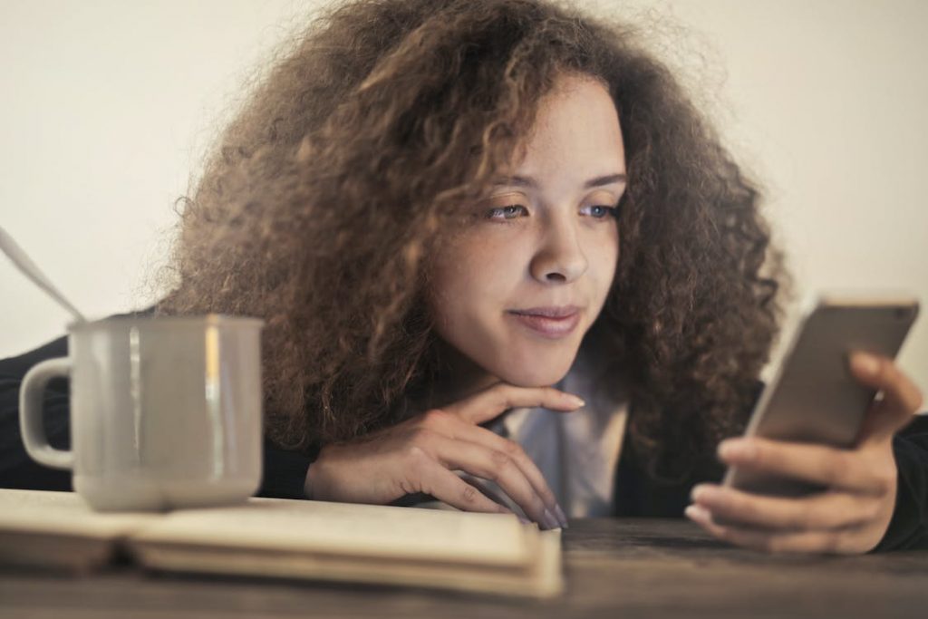 Een meisje bekijkt haar telefoon met een kop koffie en een boek naast haar. 