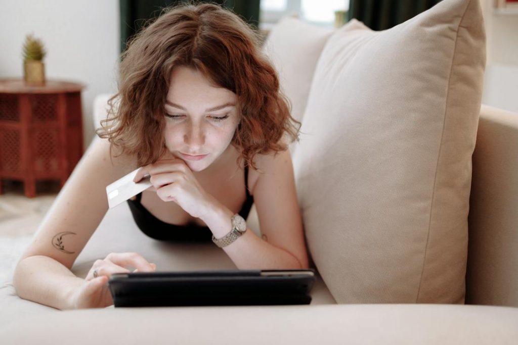 A woman on a couch using an iPad while holding a credit card on the other hand.