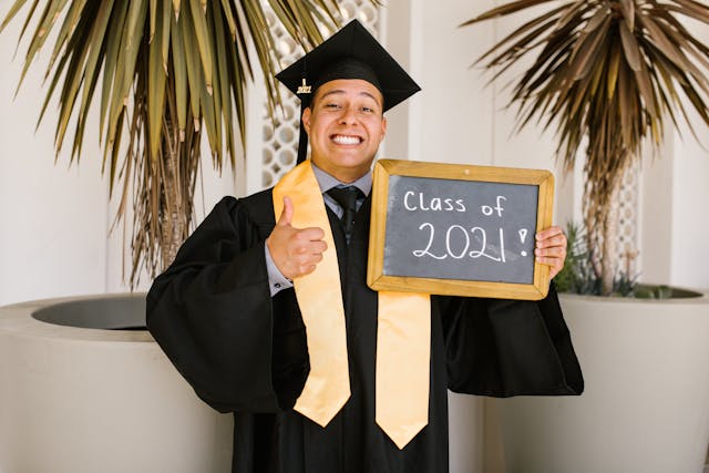 A guy wearing a toga and cap at his graduation while holding a sign that says, “Class of 2021.”