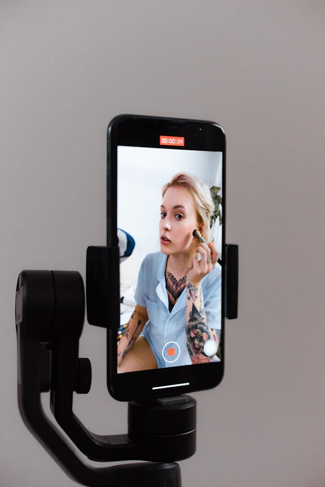 Um telemóvel com câmara num tripé a filmar uma mulher a maquilhar-se.