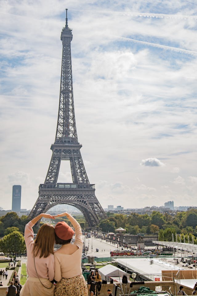 A back shot of two women looking at the Eiffel Tower while making a giant heart by connecting their arms over their heads.