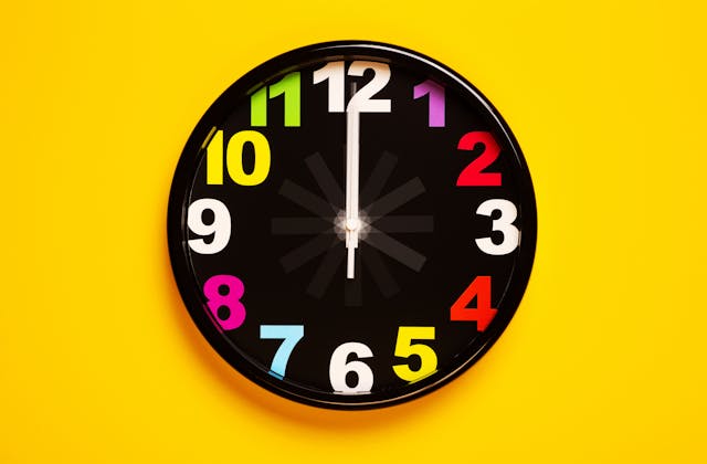 Een zwarte analoge klok met kleurrijke cijfers tegen een felgele muur.