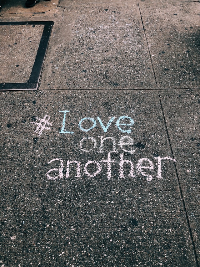 De tag #LoveOneAnother is met krijt op beton geschreven.