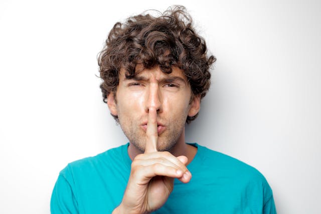 Een man in een blauw shirt met zijn vinger naar zijn lippen, maakt een sussende beweging.