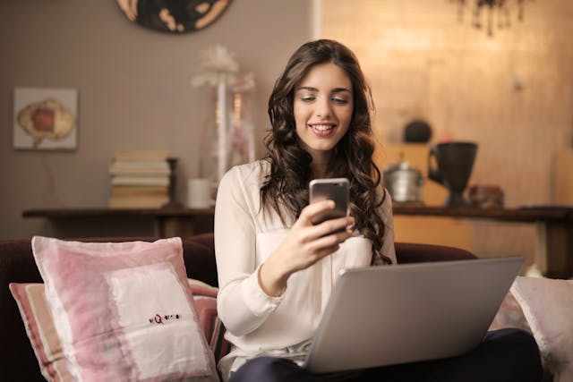 A woman smiling at her phone with a laptop on her lap.