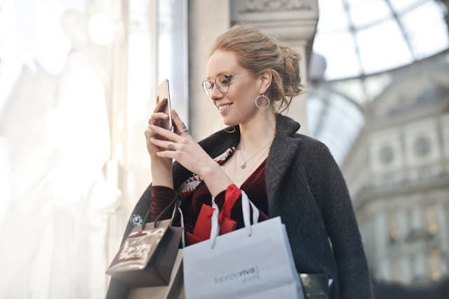 買い物袋を持ちながら、笑顔で携帯電話を見る女性。