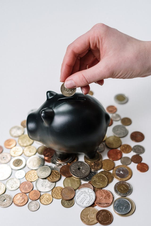 Someone putting a coin into a piggy bank with piles of other coins around it.