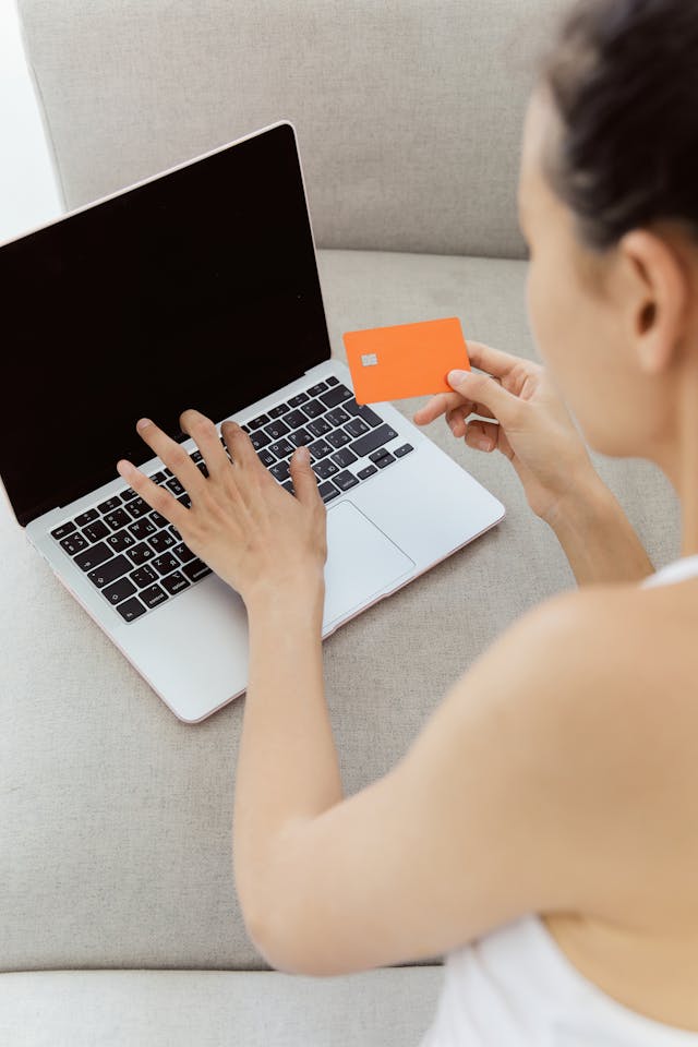 امرأة تحمل بطاقة ائتمان أثناء كتابة شيء ما على جهاز الكمبيوتر المحمول الخاص بها.