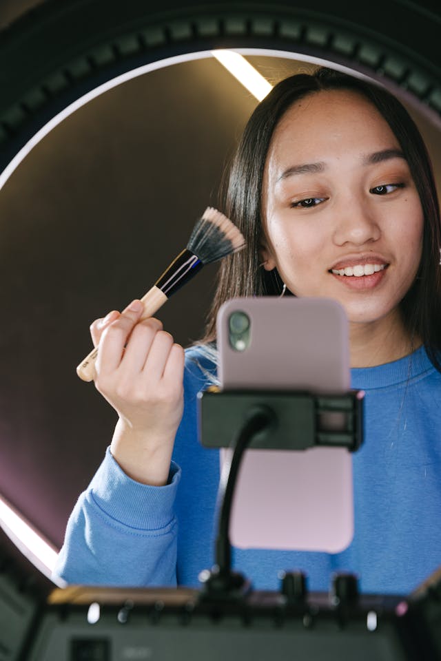 A woman doing her makeup while recording a video of herself with her phone and tripod.