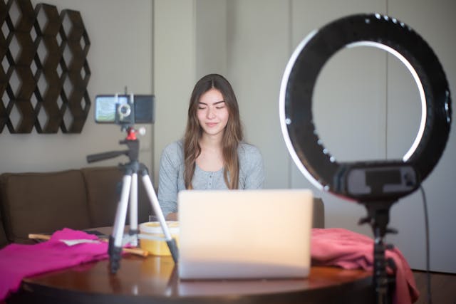A woman live-streaming with her phone, laptop, and ring light.