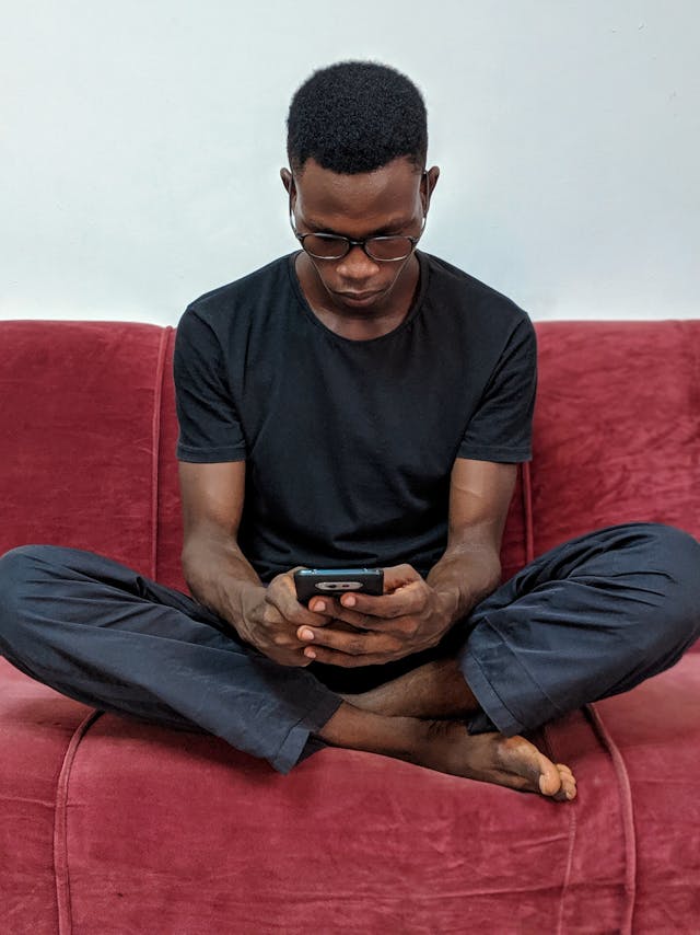 Un homme assis les jambes croisées sur un canapé tout en naviguant sur son smartphone.