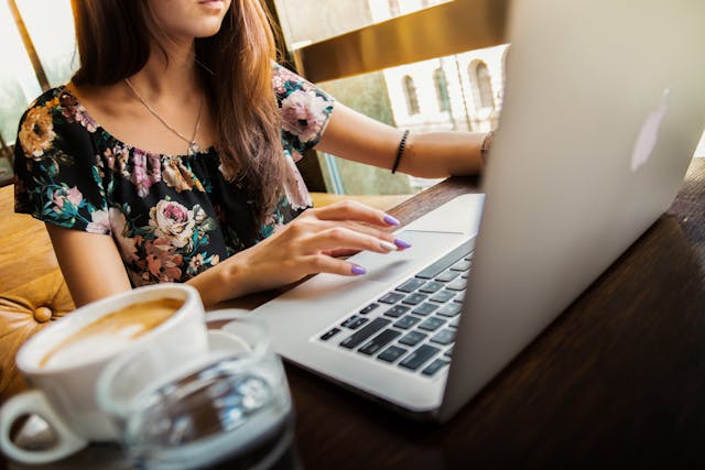 Een vrouw gebruikt haar laptop, die op een tafel staat naast haar kopje koffie.
