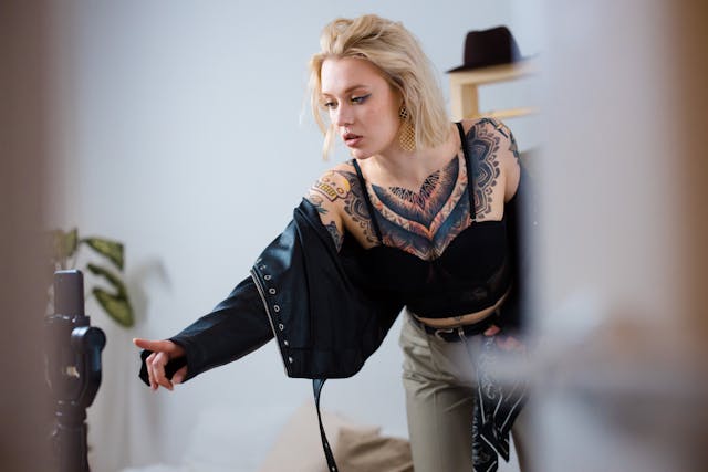 A blonde, tattooed woman recording a video of herself using her phone and tripod.