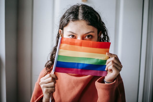 A young woman holding up an LGBT rainbow flag to cover half of her face.
