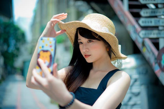 여름 모자를 쓴 여성이 휴대폰으로 귀여운 셀카를 찍고 있습니다.