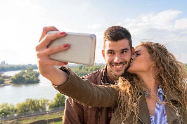 O femeie care își sărută obrazul iubitului ei în timp ce își face un selfie cu ei împreună.