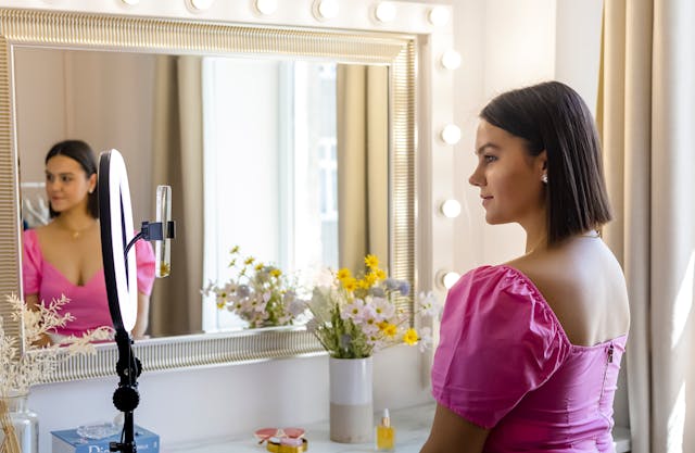 A woman sitting at her vanity filming a video of herself with her phone, ring light, and tripod.