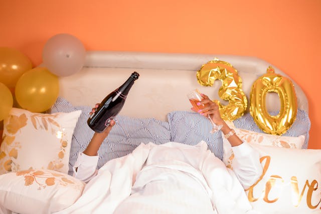 Alguém deitado debaixo das cobertas da sua cama a servir-se de uma taça de champanhe no seu 30º aniversário.