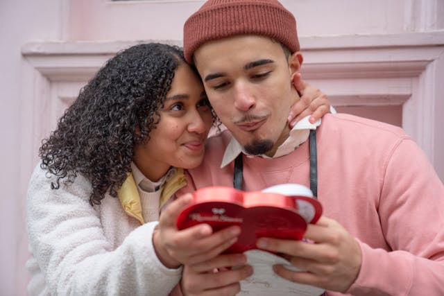 A girlfriend looking at her boyfriend as she hands him a heart-shaped box of chocolates.