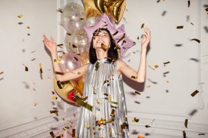 O femeie într-o rochie argintie, cu baloane în spatele ei, privește confetti aurii care cad la o petrecere aniversară.
