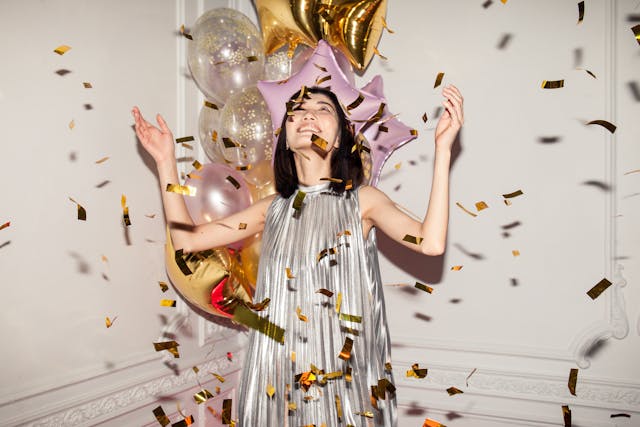 Uma mulher com um vestido prateado e balões atrás de si a olhar para os confettis dourados que caem numa festa de aniversário.