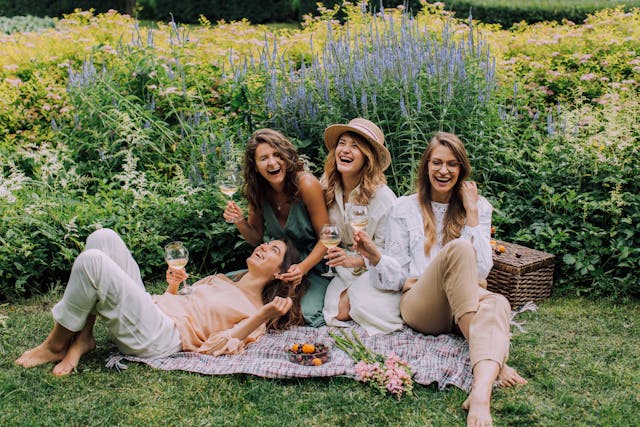 A group of female friends laughing and holding up glasses of champagne at an outdoor picnic.
