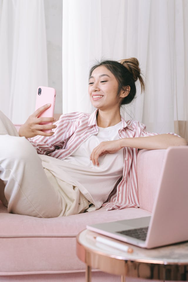 Une jeune femme assise sur un canapé rose, souriant et regardant son téléphone.