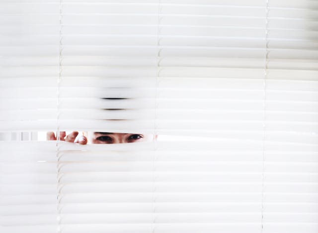 A person peeking through white window blinds.