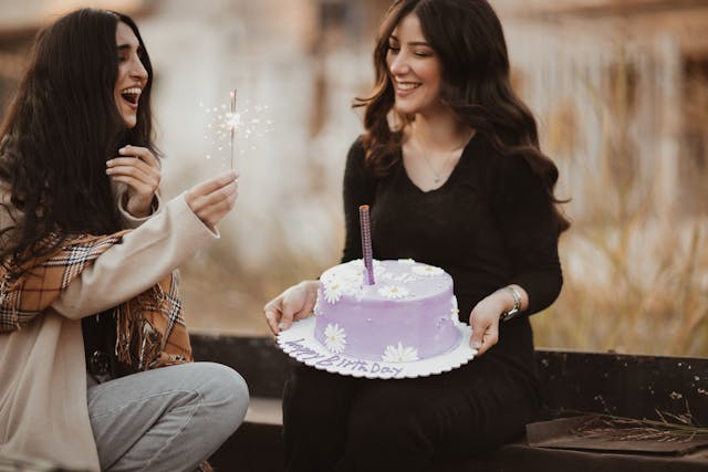 Dois amigos sentados ao ar livre, segurando um espumante e um bolo de aniversário.