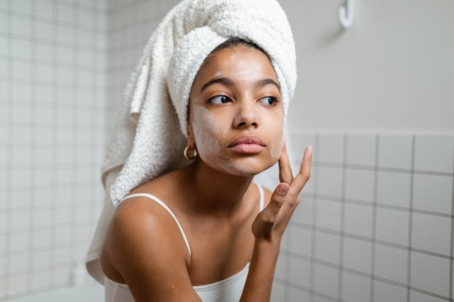 A young woman with her hair wrapped in a towel applying moisturizer on her face.