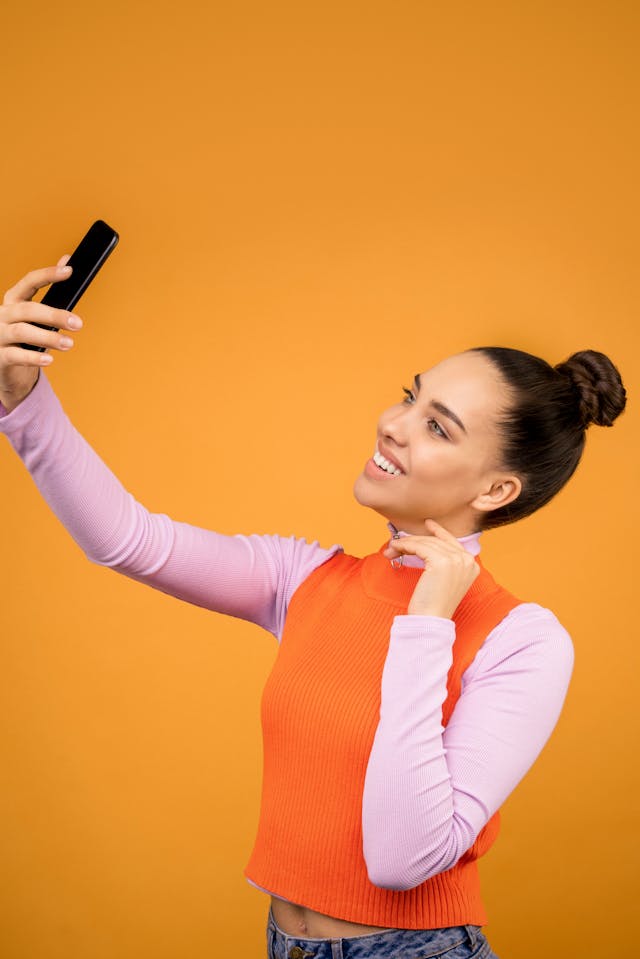 눈길을 사로잡는 주황색 배경을 배경으로 셀카를 찍기 위해 휴대폰을 들고 있는 여성.
