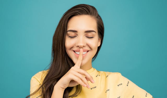 Una mujer sonriente con los ojos cerrados que se lleva un dedo a los labios para señalar un secreto.