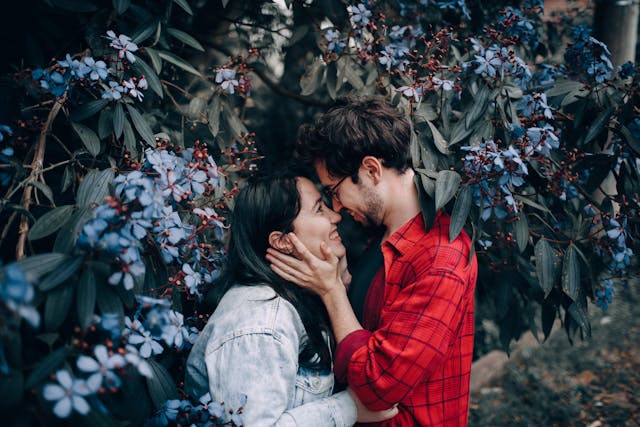 Un bărbat ținând în brațe fața iubitei sale în timp ce se uită zâmbind unul la celălalt într-o grădină.