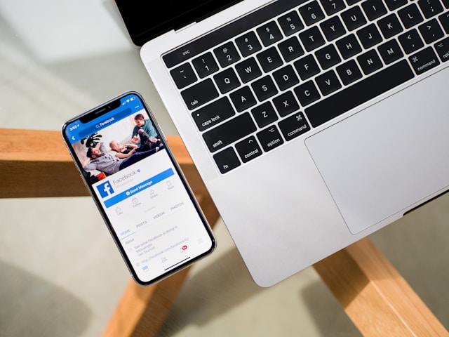 A phone displaying a Facebook page next to a laptop on a desk.