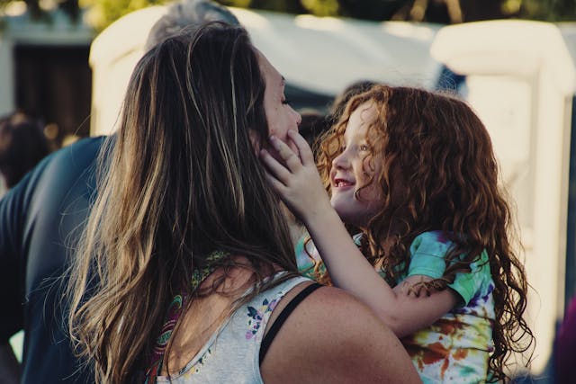 髪の乱れた小さな女の子が微笑みながら、抱っこしている母親の顔に触れている。