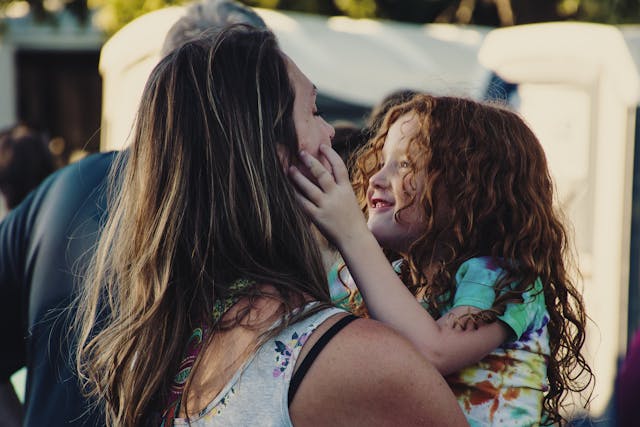 A little girl with messy hair smiling and touching her mom’s face as she carries her.