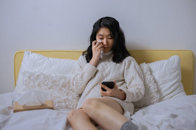 Een vrouw in bed die een traan uit haar oog veegt terwijl ze naar haar mobiele telefoon kijkt.