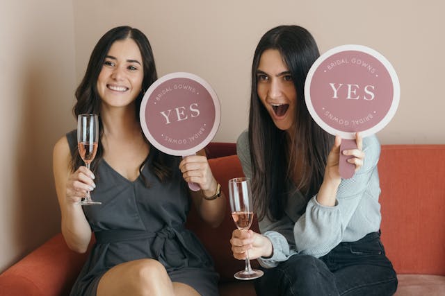 Two women holding champagne glasses and pink signs that say “Yes.”